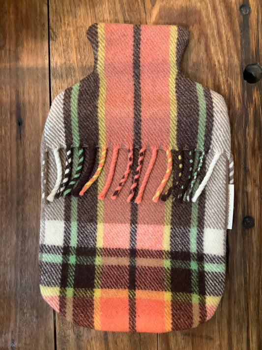 A plaid check hot water bottle cover in autumn tones and a blanket fringe sits on a wooden counter
